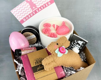 May include: A brown gift box filled with Easter-themed gifts, including a pink bunny candle, a pink lip balm, a pink soap bar, a pink egg, and a brown felt bunny. The box is decorated with pink and white shredded paper. The box also includes a card that says "Hoppy Easter".
