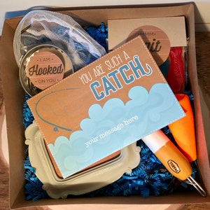 Puede incluir: Una caja de regalo llena de art&iacute;culos con tem&aacute;tica de pesca, que incluyen una red, un peque&ntilde;o frasco con una etiqueta que dice "I am Hooked on You", una tarjeta que dice "You are such a Catch", un se&ntilde;uelo de pesca y una bolsa de gusanos rojos gomosos.