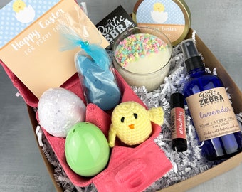 May include: A pink and white egg carton filled with Easter-themed gifts. The gifts include a lavender room spray, a bath bomb, a chick toy, a blue bunny bath bomb, a candle, and a lip balm. The carton is surrounded by white shredded paper. The card says "Happy Easter for peeps sake".