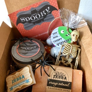 May include: A Halloween gift box filled with spooky themed items, including a jar of candle with a "Have a Spooky Halloween" label, a bar of orange oatmeal soap with a "Rustic Zebra Boutique" label, a plastic skeleton, and a bag of candy. The box also includes a card with a bat and the text "Have a Spooky Halloween".