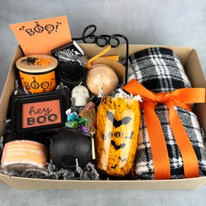 May include: A Halloween gift box containing a black and white plaid blanket with an orange ribbon, a glass with bat graphics, a black cauldron, a pumpkin souffle jar, a skull candle, a framed sign reading hey BOO, and an orange card reading BOO.