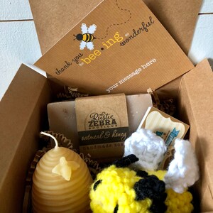 May include: A gift box filled with bee-themed items, including a handmade oatmeal and honey soap bar, a crocheted bee toy, and a honey-comb shaped candle. The box also includes a card that says "thank you for bee-ing so wonderful".