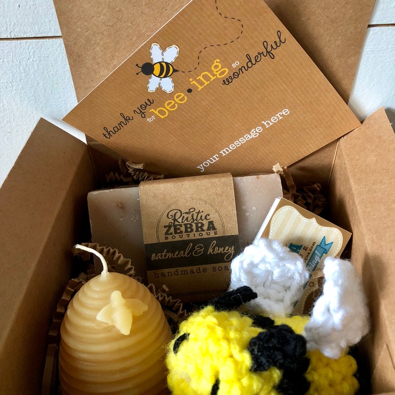 May include: A gift box filled with bee-themed items, including a handmade oatmeal and honey soap bar, a crocheted bee toy, and a honey-comb shaped candle. The box also includes a card that says "thank you for bee-ing so wonderful".