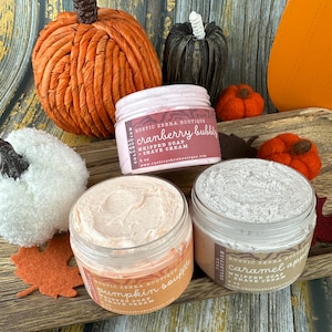 May include: An arrangement of fall-themed products, including decorative pumpkins and jars of whipped soap and shave cream. The jars are labeled with flavors like "cranberry bubbly" and "pumpkin souffl&eacute;". The image has a warm, autumnal color palette.