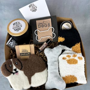 May include: A gift box filled with dog-themed items, including a brown and white felt dog toy, a white bone-shaped plush toy, a pair of white and brown fuzzy socks with paw prints, a brown wooden sign with the text "i love dogs", a small jar of bath and body products, a bar of soap, a lip balm, and a candle with a wooden lid that says "Life is better with a dog".