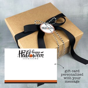 May include: A brown gift box with a black ribbon and a white and black twine bow. The box has a tag that says "Happy Halloween Countdown". The box is sitting on a gray surface. A white card with the text "Happy Halloween Countdown" and a pumpkin graphic is next to the box. The card has a black and orange border. The text "gift card personalized with your message" is written below the card.