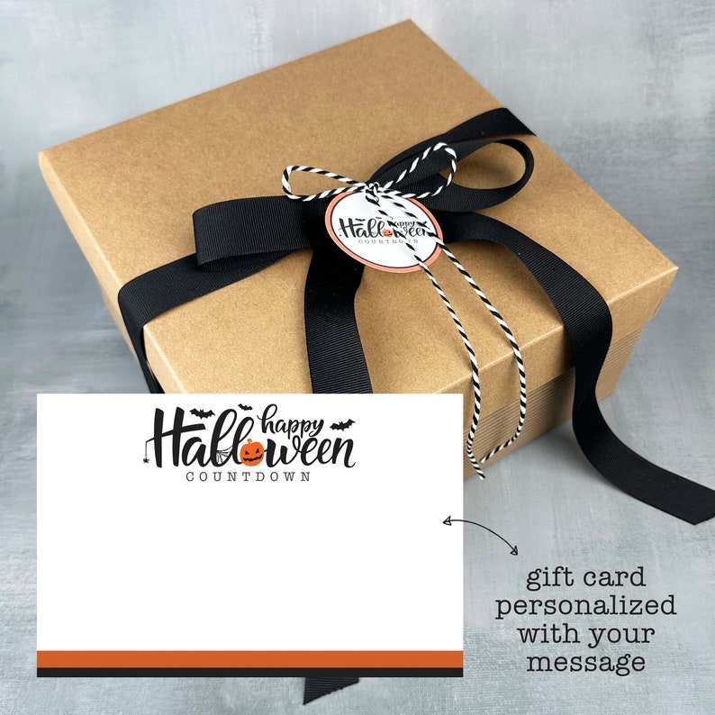May include: A brown gift box with a black ribbon and a white and black twine bow. The box has a tag that says "Happy Halloween Countdown". The box is sitting on a gray surface. A white card with the text "Happy Halloween Countdown" and a pumpkin graphic is next to the box. The card has a black and orange border. The text "gift card personalized with your message" is written below the card.