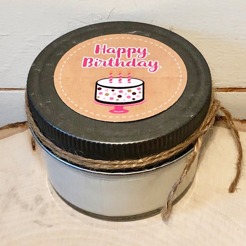 Op de afbeelding: Een klein glazen potje met een zwart metalen deksel en een bruin papieren etiket met de tekst "Happy Birthday" met een roze en bruine taart illustratie. Het potje is vastgebonden met touw.