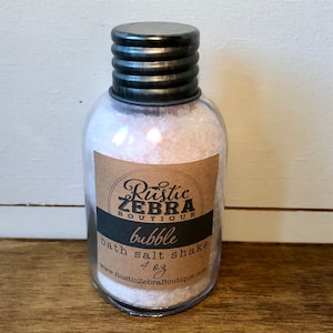 May include: A glass jar with a black metal lid filled with pink bath salts. The label reads "Rustic Zebra Boutique" and "bubble bath salt shake 4 oz" with a website address.