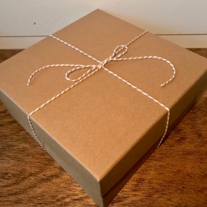 Op de afbeelding: Een bruine kartonnen doos gebonden met wit en bruin touw. De doos is ingepakt in bruin papier en heeft een eenvoudige, rustieke uitstraling.