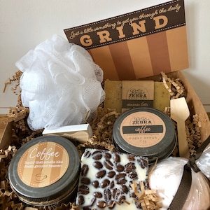 May include: A brown gift box filled with coffee-themed bath and body products. The box includes a coffee-scented sugar scrub, a coffee-scented bath bomb, a coffee-scented soap bar, a coffee-scented liquid soap, and a white bath pouf. The box also includes a wooden spoon and a card that says "Just a little something to get you through the daily GRIND".