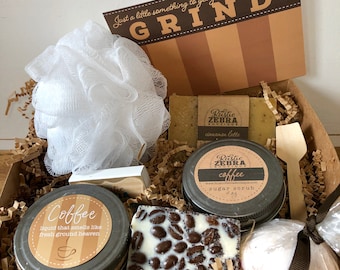 May include: A brown gift box filled with coffee-themed bath and body products. The box includes a coffee-scented sugar scrub, a coffee-scented bath bomb, a coffee-scented soap bar, a coffee-scented liquid soap, and a white bath pouf. The box also includes a wooden spoon and a card that says "Just a little something to get you through the daily GRIND".