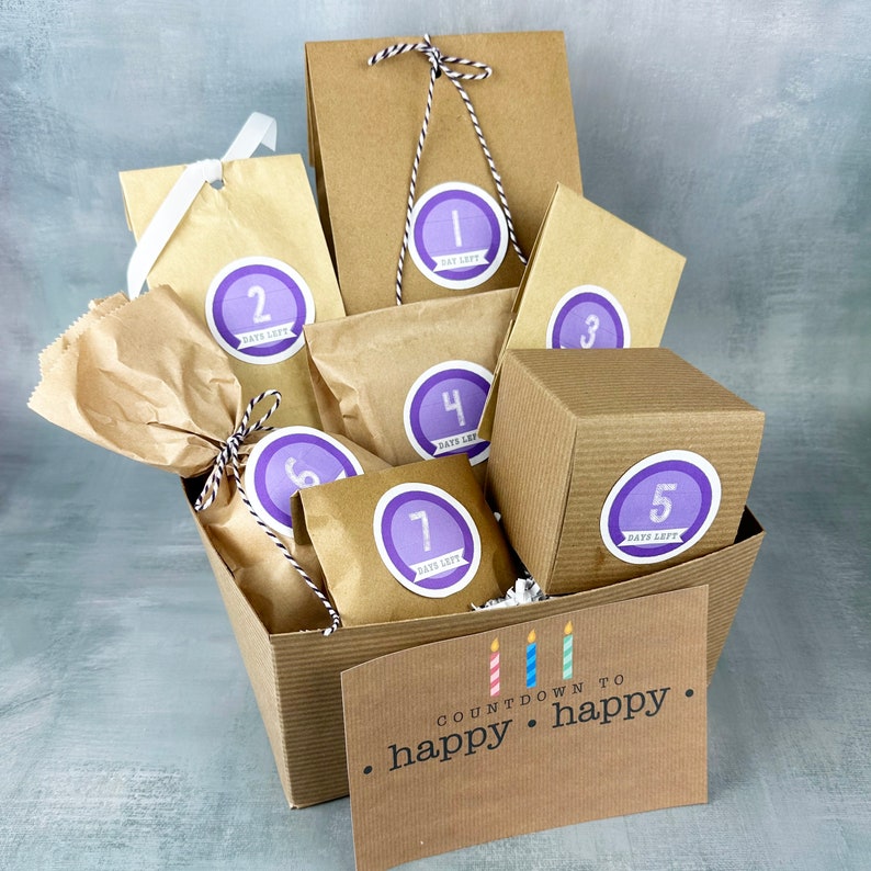 Op de afbeelding: Een bruine papieren cadeauzak gevuld met kleinere bruine papieren zakken, elk met een paarse cirkel sticker met een nummer erop. De zakken zijn gerangschikt in een mand met een bord waarop staat "COUNTDOWN TO happy happy."