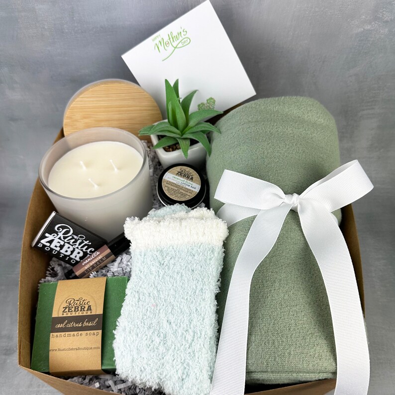 May include: A gift basket filled with self-care items. Includes a candle, a small succulent, a rolled green blanket tied with a white ribbon, a pair of fuzzy socks, a bar of soap, and a small container of lip balm.