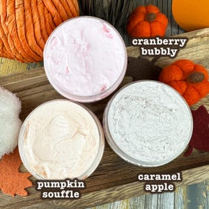 May include: Overhead shot of three open jars of body butter with the labels "cranberry bubbly", "pumpkin souffle", and "caramel apple". The jars are surrounded by fall-themed decorations, including felt pumpkins and leaves.