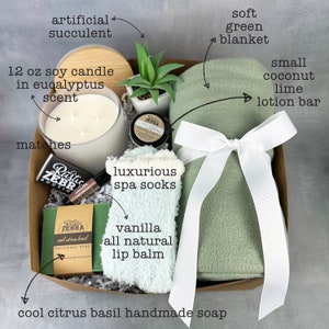 May include: A gift basket filled with self-care items. Includes a 12 oz soy candle, matches, a soft green blanket tied with a white ribbon, spa socks, lip balm, a lotion bar, handmade soap, and an artificial succulent.