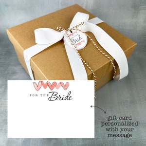 May include: A brown gift box with a white ribbon and a tag that says "For the Bride" with pink hearts. A white card with pink hearts and the text "For the Bride" is next to the box.