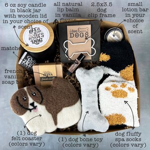 May include: A gift box filled with dog-themed items, including a 6 oz soy candle, a lip balm, a dog bone toy, a dog felt coaster, a dog clip frame, a bar of soap, and a pair of dog spa socks. The box is filled with brown shredded paper. The candle has a label that says "Life is better with a dog". The lip balm is in a small tube with a label that says "Vanilla". The dog bone toy is white with brown paw prints. The dog felt coaster is brown and white and shaped like a dog. The dog clip frame is black with a white bone-shaped frame. The bar of soap is white with a label that says "French Vanilla". The dog spa socks are white with brown paw prints. The box is decorated with a brown ribbon.