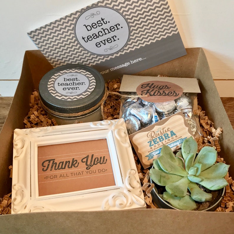 May include: A brown gift box filled with teacher appreciation gifts. The box contains a candle with a gray and white chevron label that says "best teacher ever", a small succulent plant in a tin can, a bag of chocolate kisses with a label that says "Hugs and Kisses", a small white frame with a brown card that says "Thank You - For all that you do", and a small wooden tag that says "Rustic Zebra Boutique".