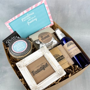 May include: A brown gift box filled with various items, including a small white frame with the text "You are Beautiful", a jar of homemade jam with a label that reads "Grandmas are moms with lots of frosting", a small bag of chocolate candies with a label that reads "Sweet", a bottle of lavender room and linen spray, and a bar of soap with a label that reads "Rustic Zebra Soap Co. -  Earl & Honey".