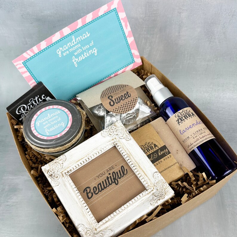 May include: A brown gift box filled with various items, including a small white frame with the text "You are Beautiful", a jar of homemade jam with a label that reads "Grandmas are moms with lots of frosting", a small bag of chocolate candies with a label that reads "Sweet", a bottle of lavender room and linen spray, and a bar of soap with a label that reads "Rustic Zebra Soap Co. -  Earl & Honey".