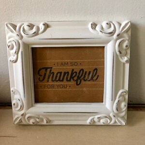 May include: A white, ornate picture frame with a distressed finish. The frame surrounds a brown background with the words "I AM SO Thankful FOR YOU" in black script. The frame has decorative corner accents.