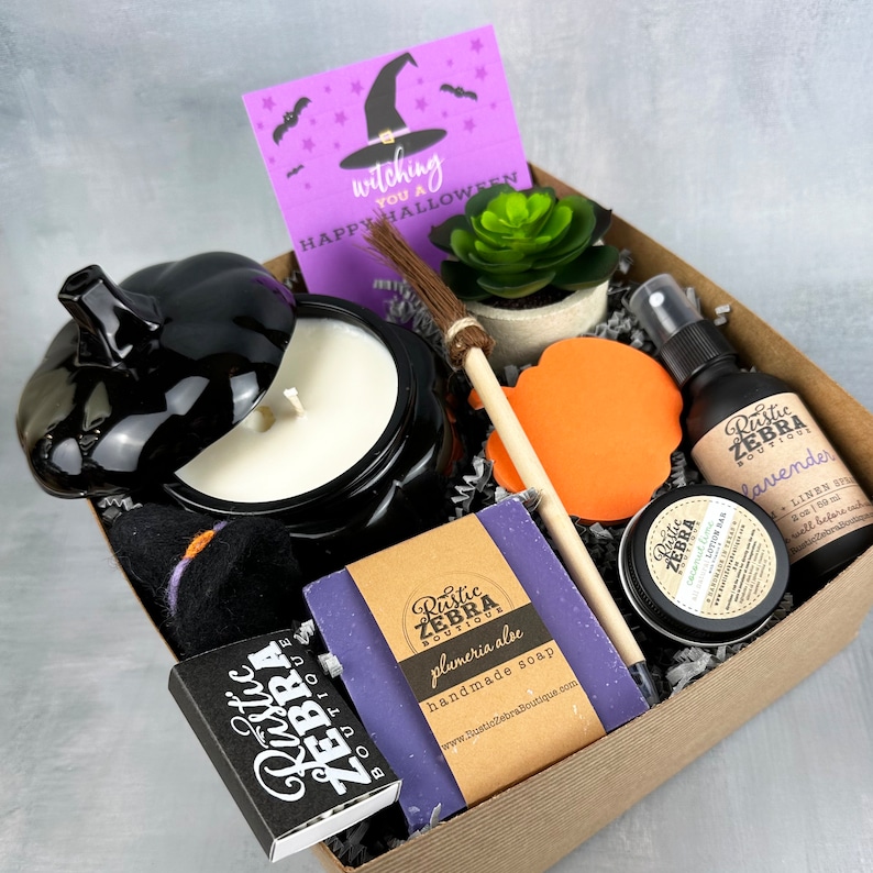 May include: A Halloween-themed gift box with a black pumpkin-shaped candle, a purple soap bar, and a lavender linen spray. The box includes a card that says "witching you a Happy Halloween". Other items include a small broom, a bath bomb, and a succulent.