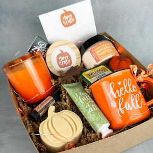 May include: A fall-themed gift box filled with an orange candle, a mug with "hello fall" text, a pumpkin-shaped soap, lip balm, lotion, and a card with a pumpkin graphic. The box includes a small pumpkin decoration and tea.