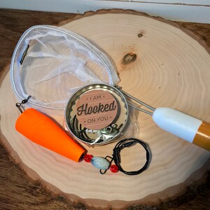 Puede incluir: Un juego de regalo con tem&aacute;tica de pesca con una red peque&ntilde;a, un se&ntilde;uelo, un flotador y una lata redonda con una etiqueta que dice "Estoy enganchado a ti".