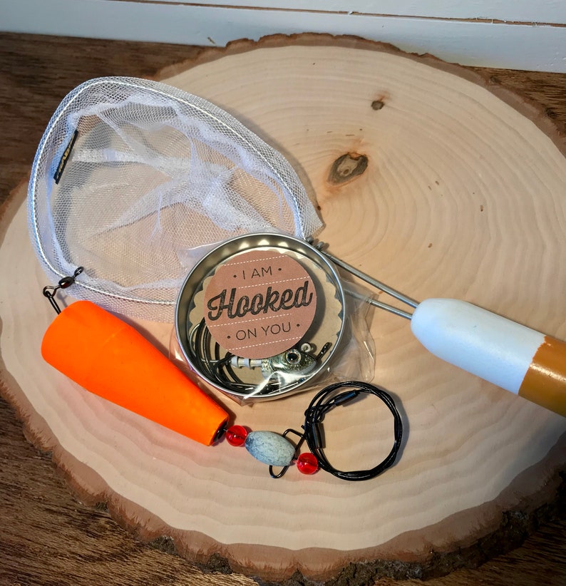 Puede incluir: Un juego de regalo con tem&aacute;tica de pesca con una red peque&ntilde;a, un se&ntilde;uelo, un flotador y una lata redonda con una etiqueta que dice "Estoy enganchado a ti".
