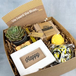 May include: A gift box filled with a variety of items, including a succulent plant, a small picture frame with the text "BEE Happy", a bar of soap, a candle, and a small tin with a bee design. The box is decorated with brown paper and yellow tissue paper.