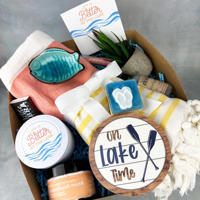 May include: A gift basket filled with lake-themed items. Includes a ceramic fish, a towel, a candle, a soap, a lip balm, a small plant, and a sign that says "On Lake Time". The card reads "Life is Better at the Lake".