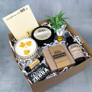 May include: A brown gift box filled with a variety of bee-themed bath and body products, including a candle, soap, lotion bar, lip balm, and linen spray. The box is filled with shredded paper and includes a small potted succulent plant. The box also includes a handwritten note that says "Everything will BEE ok".