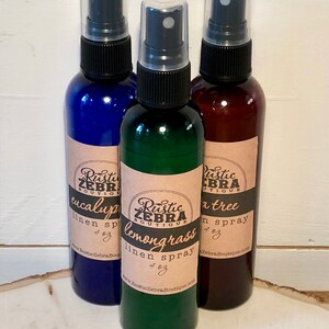 May include: Three linen spray bottles in blue, green, and brown. Each bottle has a black spray nozzle and a label with the text "Rustic Zebra Boutique" and the scent name: eucalyptus, lemongrass, and tea tree. Each bottle is 4 oz.