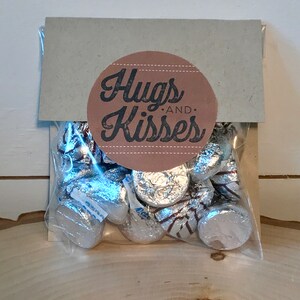 May include: A brown paper bag with a round sticker that says "Hugs and Kisses" in black text. The bag contains silver foil-wrapped chocolate kisses.