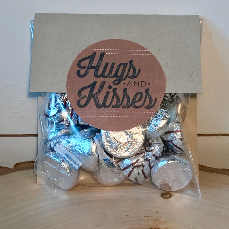 May include: A brown paper bag with a round sticker that says "Hugs and Kisses" in black text. The bag contains silver foil-wrapped chocolate kisses.