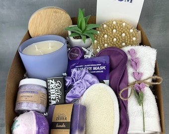 May include: A gift basket filled with lavender-themed bath and body products, including a candle, bath salts, a bath bomb, soap, a sleep mask, a hair scrunchie, and a loofah. The basket also includes a small potted succulent plant and a card that says "I love you Mom".
