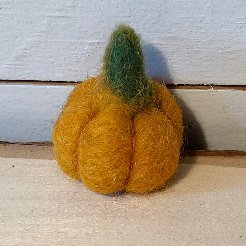 May include: A small, handmade pumpkin made of orange felt with a green stem. The pumpkin has a ribbed texture and is sitting on a light-colored wooden surface. A seasonal autumn decoration.