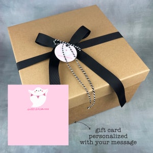 May include: A brown gift box with a black ribbon and a white tag that says "Happy Halloween". A pink card with a ghost holding a banner that says "BOO" is in the foreground. The card says "Happy Halloween".