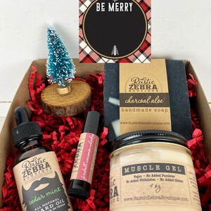 May include: A gift box filled with a variety of men's grooming products, including beard oil, lip balm, soap, and muscle gel. The box is decorated with red and white tissue paper and a small Christmas tree. The products are all from the Rustic Zebra Boutique and feature a rustic, masculine design.