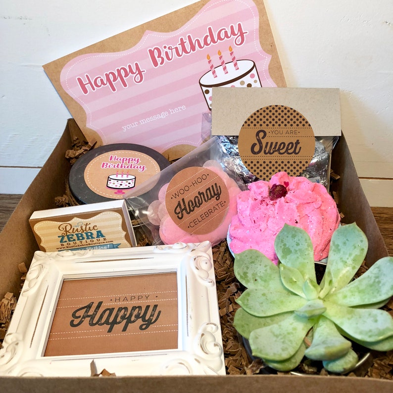 Op de afbeelding: Een bruine cadeaudoos gevuld met cadeaus met een verjaardags thema. De doos bevat een roze badbom, een kleine vetplant, een ingelijst bord met de tekst "Happy", een kleine kaars met een verjaardagstaartontwerp, een klein bord met de tekst "You are Sweet" en een klein bord met de tekst "Woo-hoo Hooray Celebrate".