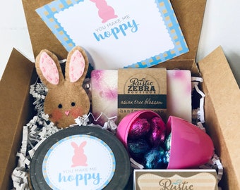 May include: A gift box filled with Easter-themed items, including a brown felt bunny, a pink egg filled with chocolate candies, a jar of homemade candle with a bunny and "You make me hoppy" label, and two bars of soap with a "Rustic Zebra Boutique" label.