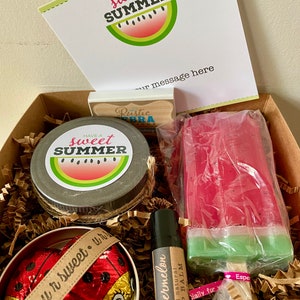 May include: A gift box filled with summer-themed items, including a watermelon-scented lip balm, a watermelon-shaped soap, a jar of hand cream, and chocolate ladybug candies. The box is lined with brown paper shreds and includes a card that says "Have a sweet summer".