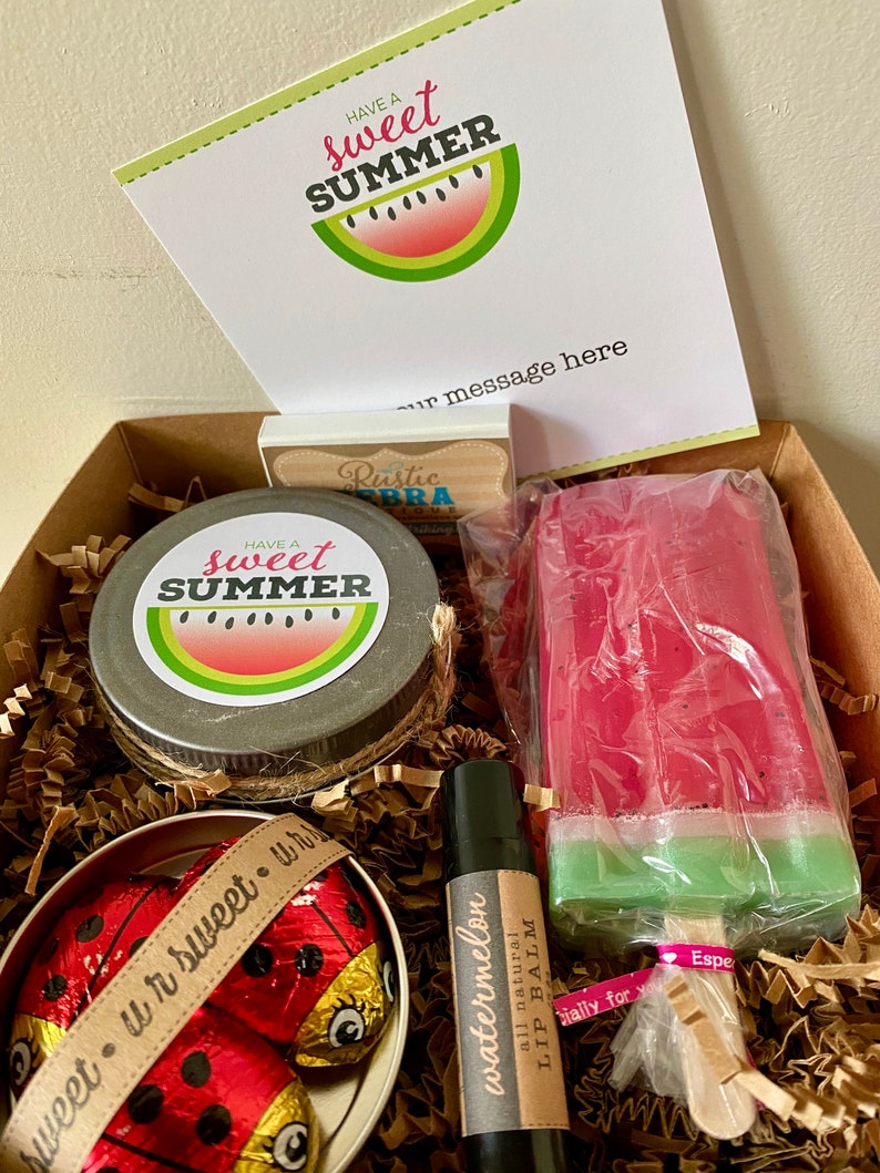 May include: A gift box filled with summer-themed items, including a watermelon-scented lip balm, a watermelon-shaped soap, a jar of hand cream, and chocolate ladybug candies. The box is lined with brown paper shreds and includes a card that says "Have a sweet summer".