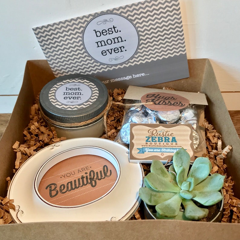 May include: A gift box filled with brown shredded paper and a variety of items, including a candle with a label that says "best mom ever", a small plate with the words "You are Beautiful", a bag of chocolate kisses, a succulent plant in a small pot, and a card with the words "best mom ever" and a space for a message.