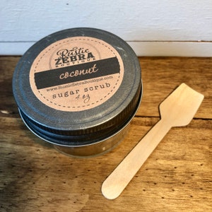 May include: A small glass jar with a black lid and a brown label that reads "Rustic Zebra Boutique" and "coconut sugar scrub 4 oz"  The jar is sitting on a wooden surface with a wooden spoon next to it.