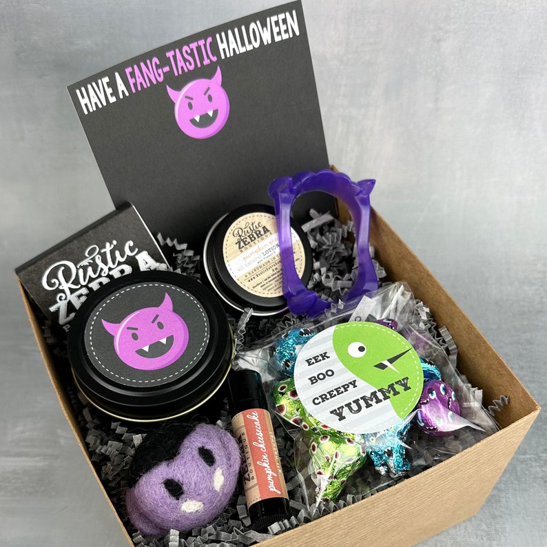May include: A Halloween-themed gift box filled with various items. Includes a black card with "HAVE A FANG-TASTIC HALLOWEEN" text, a purple devil graphic, and a felt vampire. Also contains a lip balm, lotion, and candies.