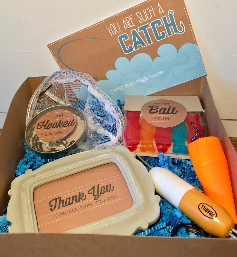 Puede incluir: Una caja de cart&oacute;n marr&oacute;n llena de regalos con tem&aacute;tica de pesca. La caja contiene un peque&ntilde;o marco blanco con el texto "Gracias. Por todo lo que haces."  Un peque&ntilde;o recipiente de metal con una etiqueta blanca que dice "Estoy enganchado a ti."  Una peque&ntilde;a bolsa de peces de goma con una etiqueta que dice "Mmm Bait Yum."  Un se&ntilde;uelo de pesca blanco y naranja y una peque&ntilde;a red.
