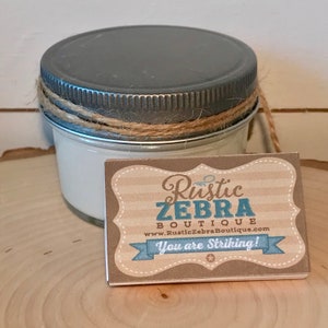 May include: A small glass jar candle with a gray lid and a brown twine bow. The candle is labeled "Rustic Zebra Boutique" with the website address "www.RusticZebraBoutique.com" and the text "You are Striking!"