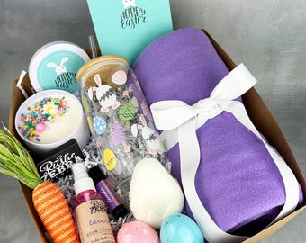 May include: A gift basket filled with Easter-themed items, including a purple throw blanket, a glass tumbler with a bunny and egg design, a candle, bath bombs, a carrot, and a bottle of lavender essential oil. The basket is tied with a white ribbon and includes a card that says "Happy Easter".
